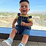 blue_sky, casual_clothing, cheeks, child, cute, denim_overalls, fashion, floor, gold_necklace, haircut, happy, indoor, natural_light, person, portrait, shorts, smiling, sneakers, toddler, window