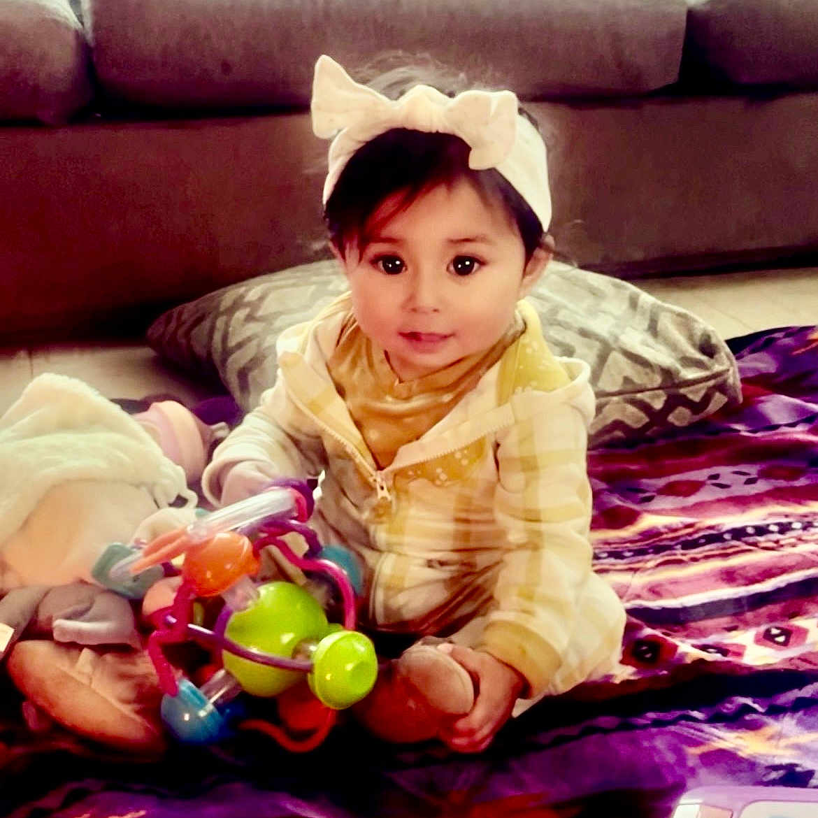 Enveeluna is registered to the contest to win money with this photo: baby, blanket, child, clothing, colorful, cushion, cute, face, floor, headband, home, indoor, infant, person, play, plush_toy, sitting, smile, toy, warm_lighting