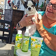 Shana participe au concours pour gagner de l'argent avec cette photo : dog, person, skincare_products, table, outdoor, balcony, glass_table, bottle, tube, jar, lemon, decor, rooster_statue, woman, glasses, smile, hand, sunlight, casual_clothing, pet