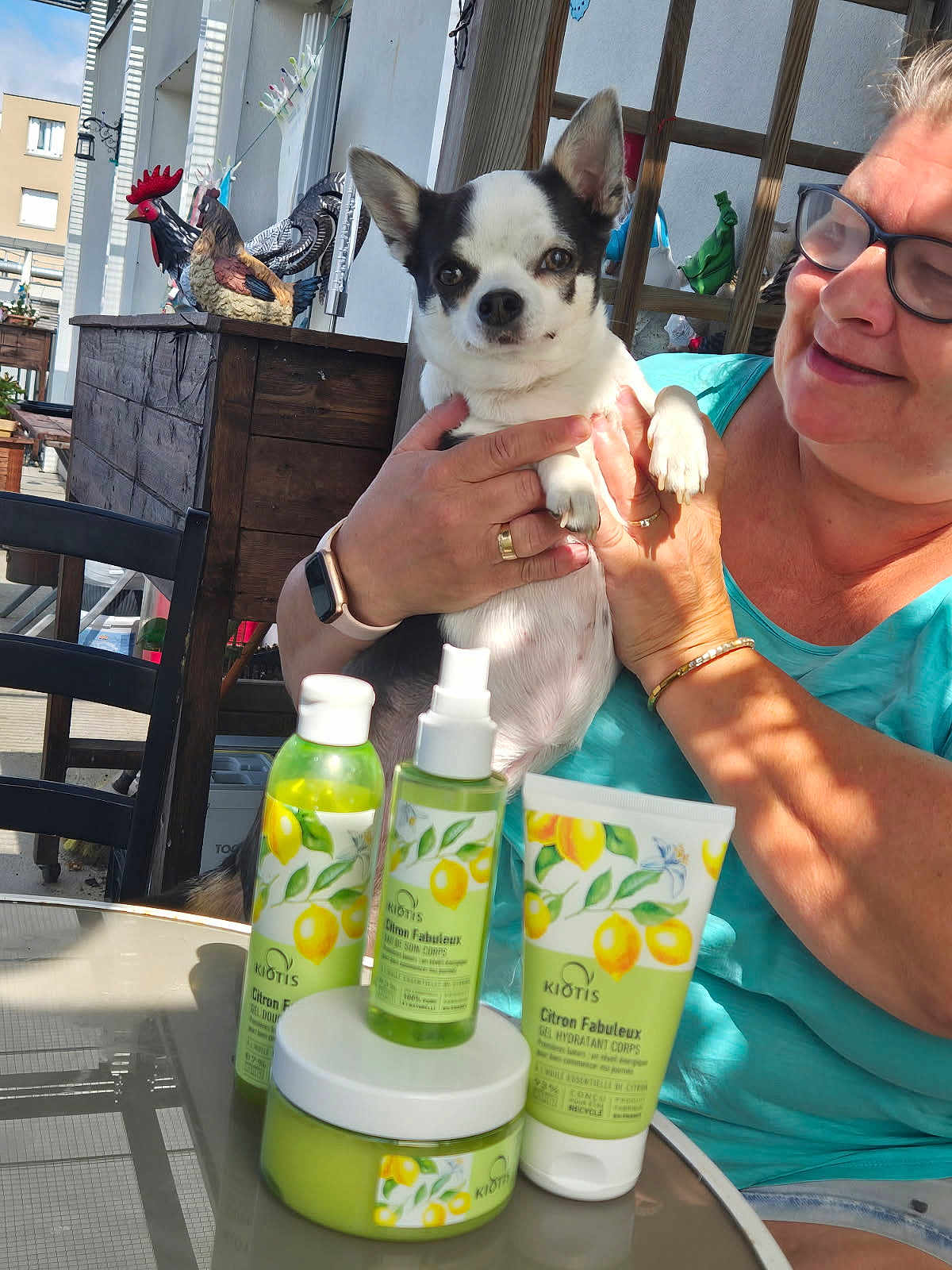 Shana participe au concours pour gagner de l'argent avec cette photo : dog, person, skincare_products, table, outdoor, balcony, glass_table, bottle, tube, jar, lemon, decor, rooster_statue, woman, glasses, smile, hand, sunlight, casual_clothing, pet