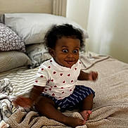 Amina is registered to the contest to win money with this photo: baby, child, bed, blanket, pillow, clothing, heart_pattern, striped_shorts, indoor, person, cute, smiling, sitting, soft_texture, room, infant, happy, cozy, dark_skin, curly_hair