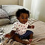 baby, child, bed, blanket, pillow, clothing, heart_pattern, striped_shorts, indoor, person, cute, smiling, sitting, soft_texture, room, infant, happy, cozy, dark_skin, curly_hair