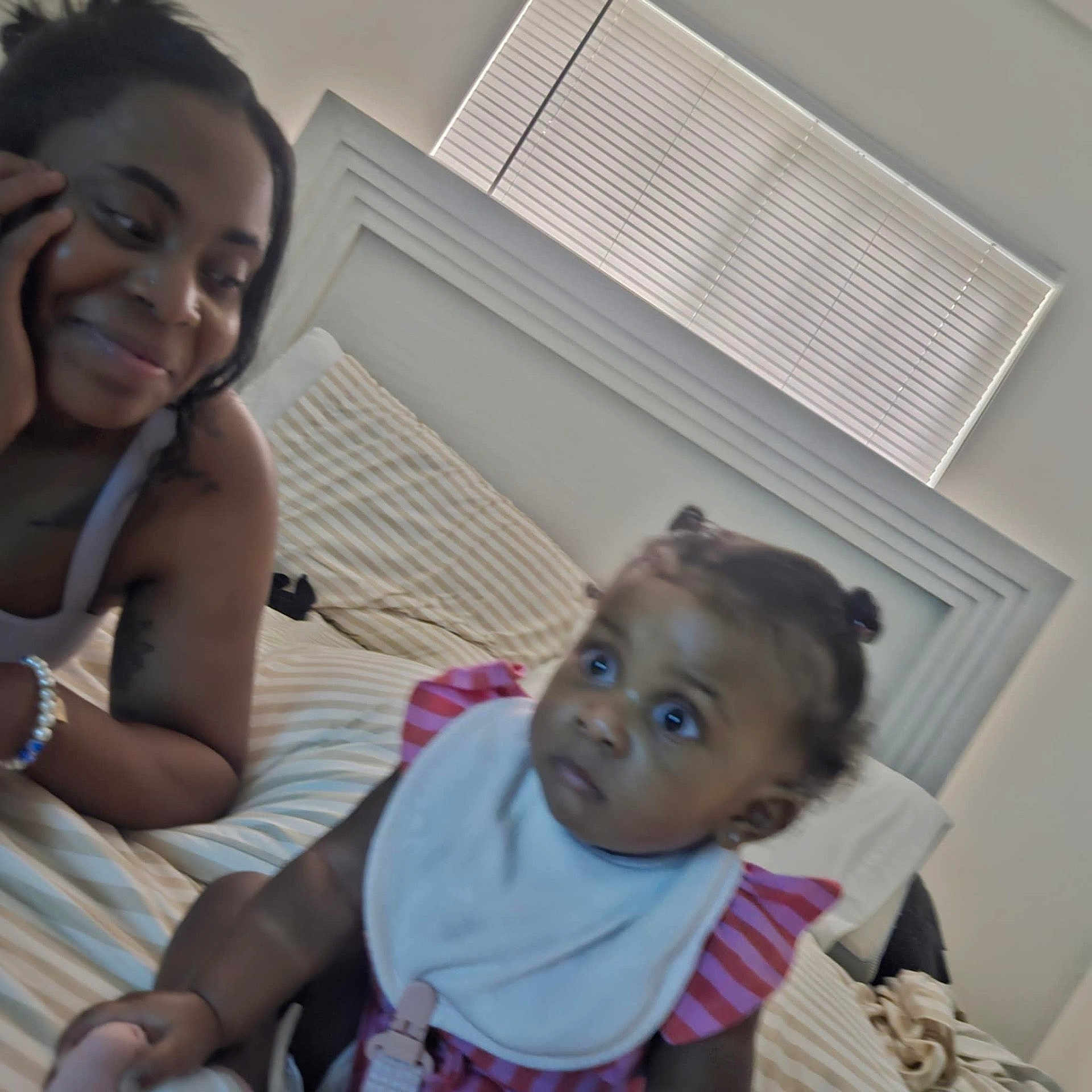 Amina is registered to the contest to win money with this photo: baby, woman, bed, indoor, smile, person, child, hair, pigtails, bib, striped_clothing, window, headboard, blanket, bracelet, skin, face, expression, interaction, family