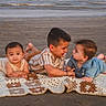 child, baby, toddler, blanket, beach, sand, ocean, water, waves, lying_down, curious, smiling, interaction, sunset, outdoor, casual_clothing, family, siblings, playful, crocheted_blanket