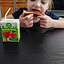child, bread, juice_box, table, food, snack, kitchen, furniture, indoors, person, eating, brown_hair, wood, cabinet, television, black_table, casual, young, girl, happy