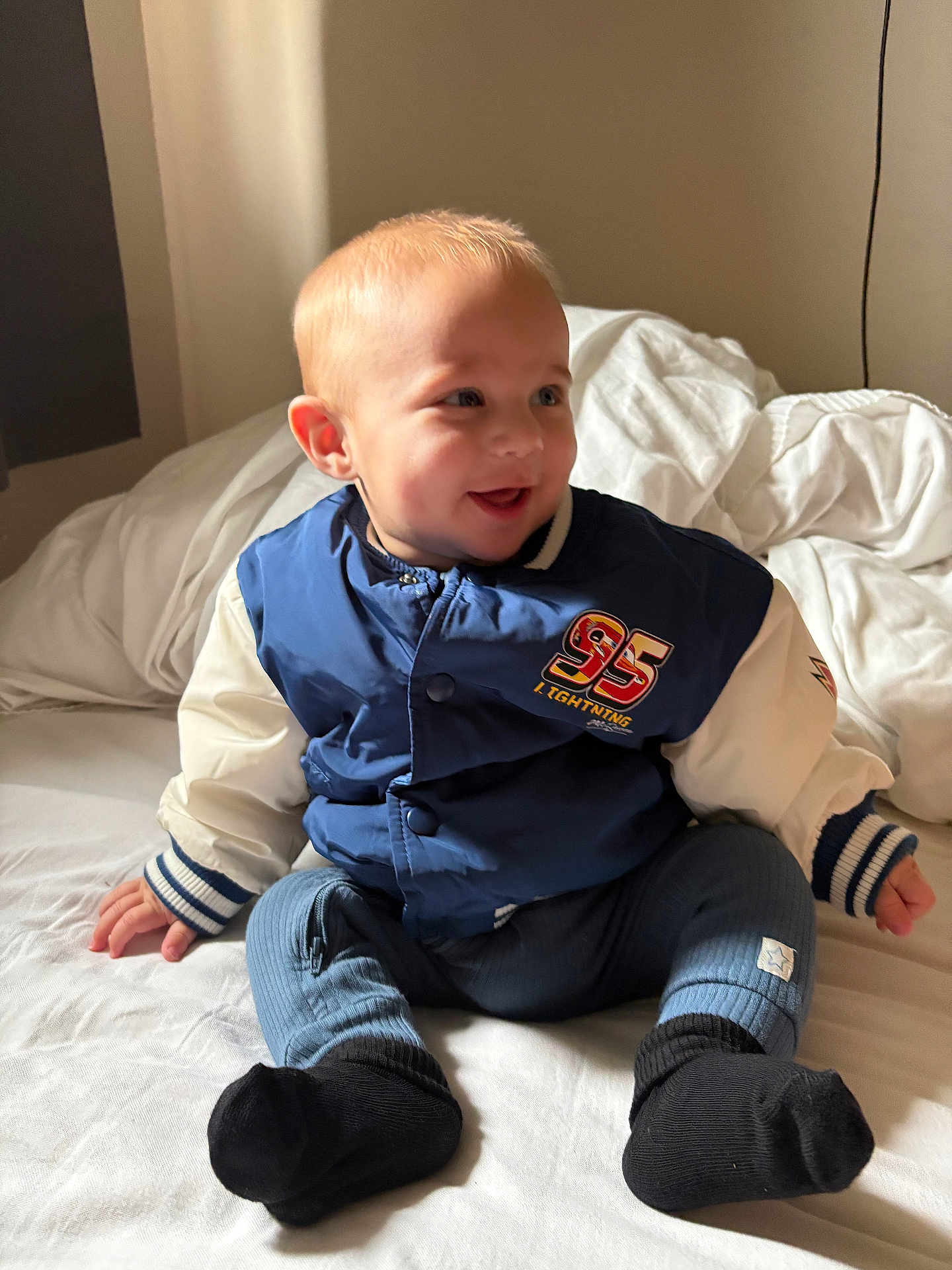 Louie joined the competition — help win amazing prizes! baby, child, infant, smiling, sitting, bed, jacket, blue_jacket, black_socks, white_bed, indoor, natural_light, cute, happy, portrait, clothing, soft_light, blonde_hair, person, young_child