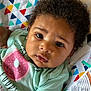 baby, infant, child, curly_hair, face, eyes, expression, mint_green_shirt, donut_graphic, colorful_blanket, patterned_blanket, close_up, lying_down, cute, adorable, skin, earrings, soft_lighting, indoors, portrait