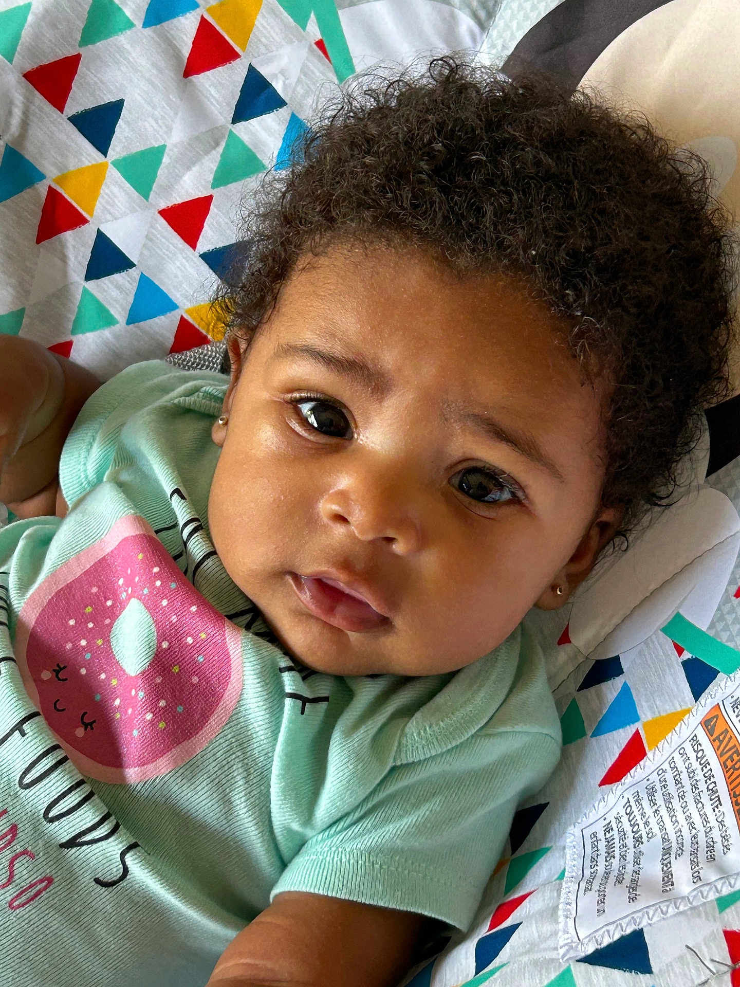 Bremi joined the competition — help win amazing prizes! baby, infant, child, curly_hair, face, eyes, expression, mint_green_shirt, donut_graphic, colorful_blanket, patterned_blanket, close_up, lying_down, cute, adorable, skin, earrings, soft_lighting, indoors, portrait