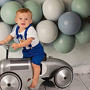 Ryder joined the competition — help win amazing prizes! toddler, child, toy_car, blue_suspender, white_tshirt, balloons, pastel_colors, indoor, flooring, barefoot, steering_wheel, portrait, cute, happy, smiling, blonde_hair, blue_eyes, playful, background, studio