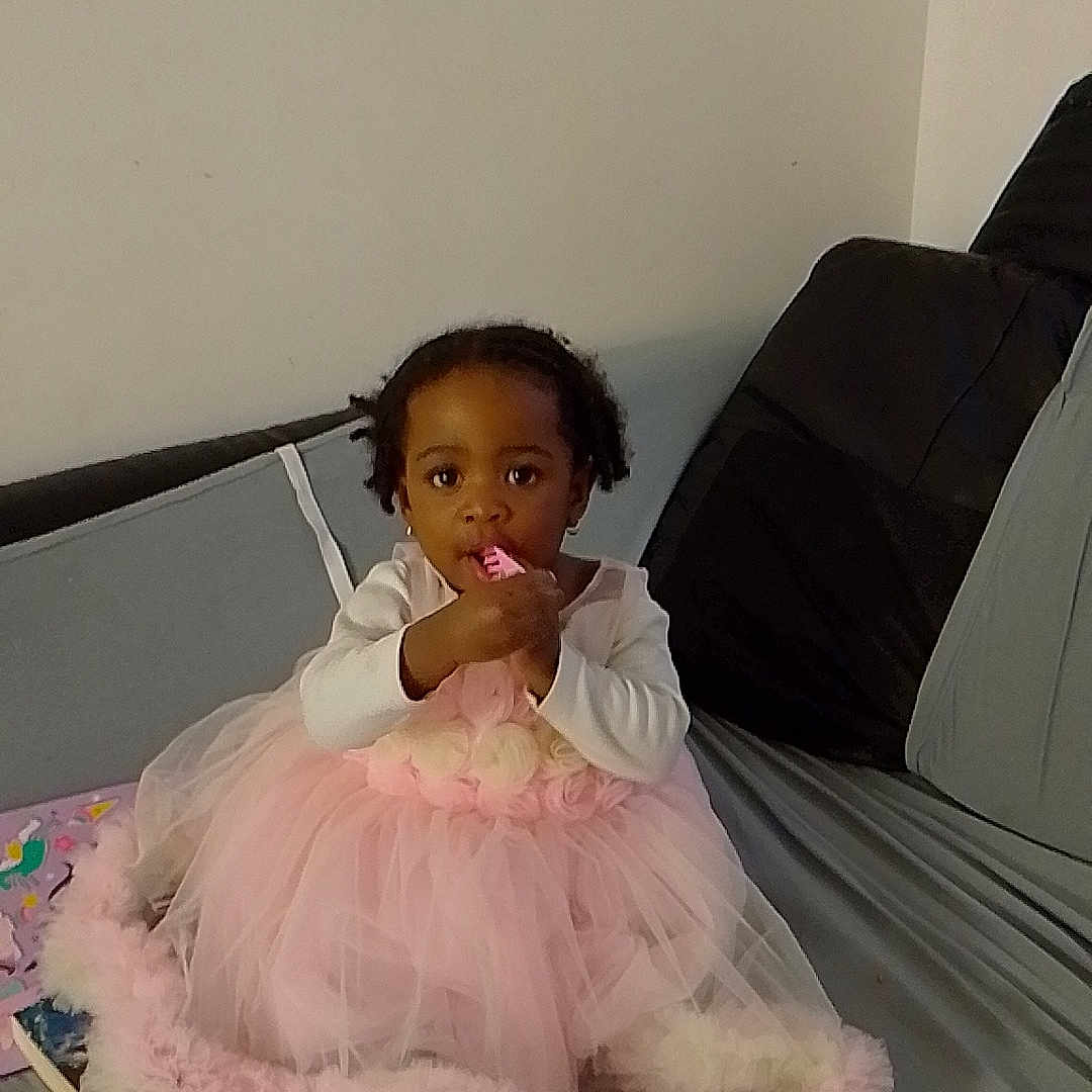 Theresa is registered to the contest to win money with this photo: barefoot, book, child, clothing, couch, cute, dress, face, girl, hair, holding, home, indoor, person, pink, playful, sitting, toddler, toy, young_child