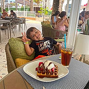 Luca participe au concours pour gagner de l'argent avec cette photo : child, boy, smiling, peace_sign, waffle, ice_cream, strawberries, chocolate_sauce, iced_tea, glass, straw, chair, cushion, table, outdoor_cafe, people, patio, palm_tree, sunlight, relaxation