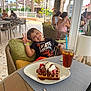 child, boy, smiling, peace_sign, waffle, ice_cream, strawberries, chocolate_sauce, iced_tea, glass, straw, chair, cushion, table, outdoor_cafe, people, patio, palm_tree, sunlight, relaxation