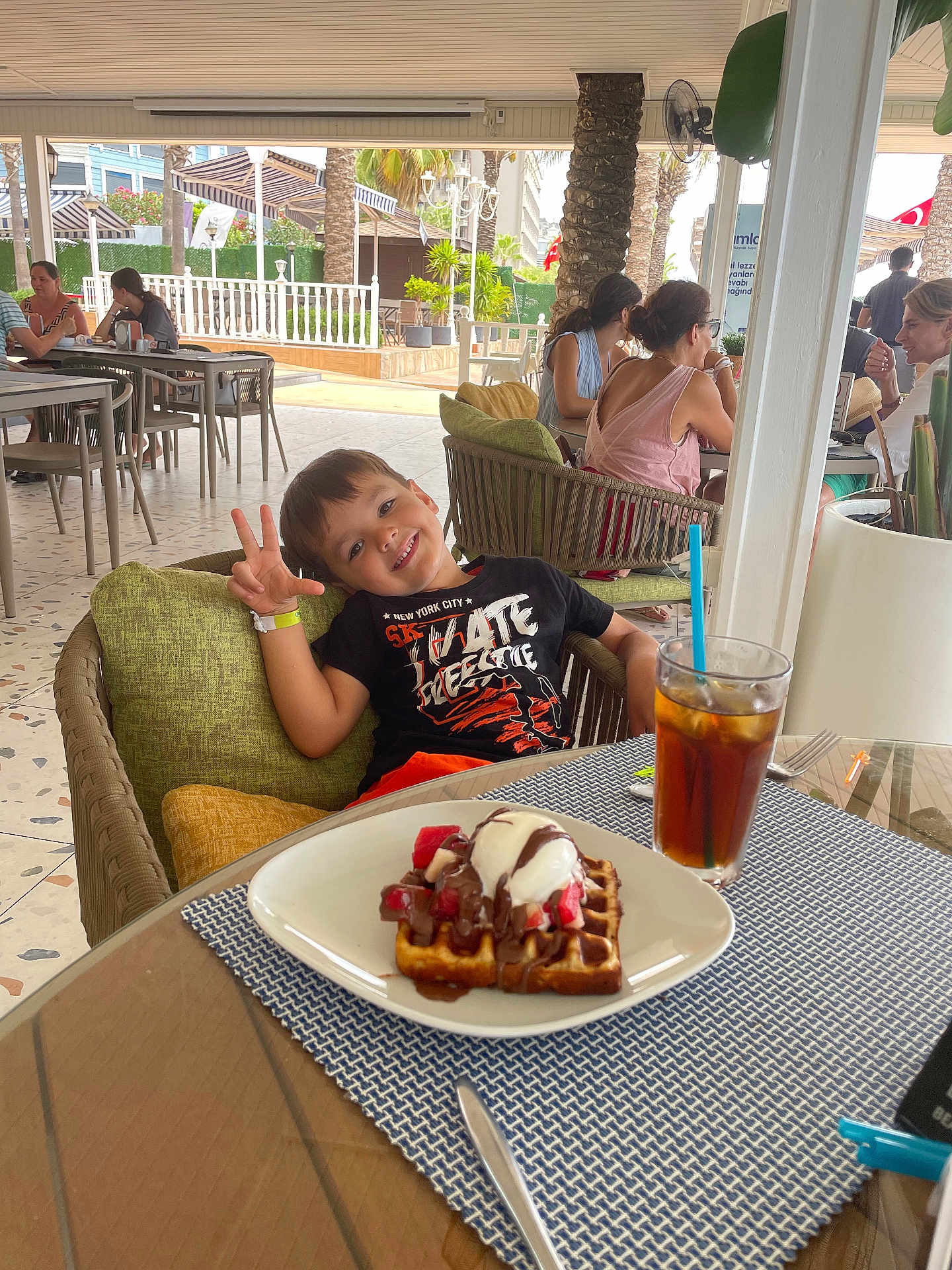 Luca participe au concours pour gagner de l'argent avec cette photo : child, boy, smiling, peace_sign, waffle, ice_cream, strawberries, chocolate_sauce, iced_tea, glass, straw, chair, cushion, table, outdoor_cafe, people, patio, palm_tree, sunlight, relaxation