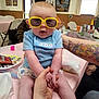Stella is registered to the contest to win money with this photo: baby, sunglasses, blue_shirt, table, hand, indoor, person, food, snack, chair, tattoo, arm, window, curtain, plate, diaper, pink_tablecloth, expression, holding_hands, casual