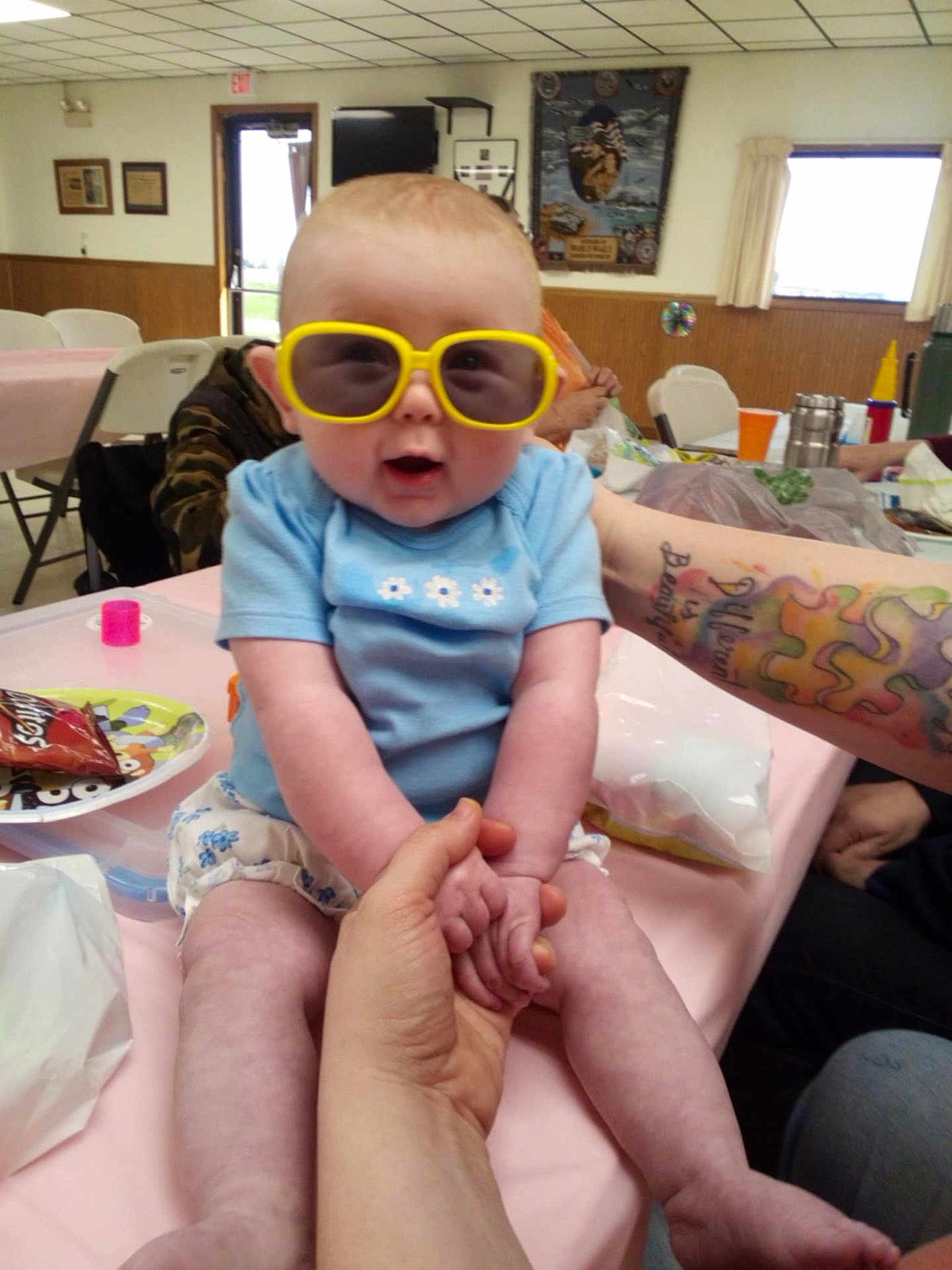 Stella is registered to the contest to win money with this photo: baby, sunglasses, blue_shirt, table, hand, indoor, person, food, snack, chair, tattoo, arm, window, curtain, plate, diaper, pink_tablecloth, expression, holding_hands, casual
