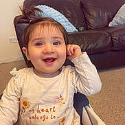 Inaya joined the competition — help win amazing prizes! toddler, child, smiling, high_chair, indoor, living_room, leather_couch, pillow, carpet, long_sleeves, hair_tie, cute, happy, portrait, baby, playful, sitting, clothing, face, person