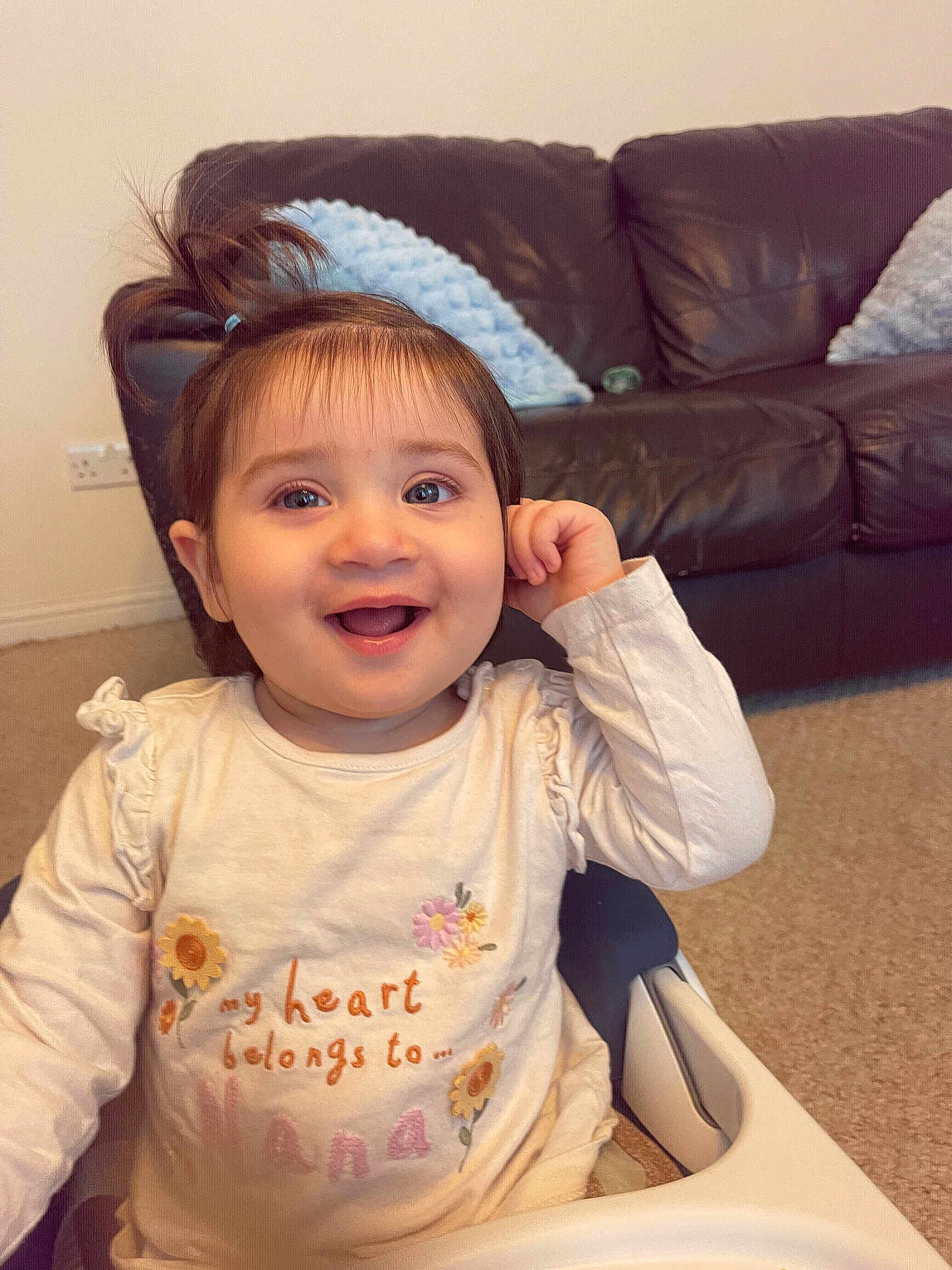 Inaya joined the competition — help win amazing prizes! toddler, child, smiling, high_chair, indoor, living_room, leather_couch, pillow, carpet, long_sleeves, hair_tie, cute, happy, portrait, baby, playful, sitting, clothing, face, person