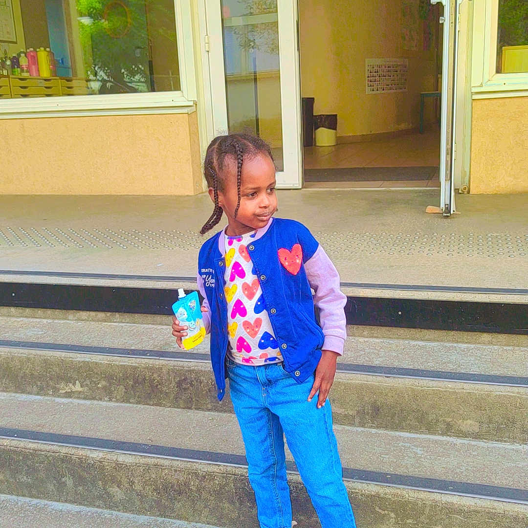 Bara'A a rejoint le concours — aidez-le/la à gagner de superbes lots ! blue_jacket, building, casual_clothing, child, concrete, confident_pose, daylight, door, drink_pouch, glass, hair_braids, heart_pattern, jeans, outdoor, person, reflection, sneakers, standing, steps, urban