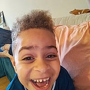 Eliott a rejoint le concours — aidez-le/la à gagner de superbes lots ! child, smiling, laughing, curly_hair, blue_shirt, couch, cushion, plush_toy, living_room, happy, face, person, indoors, closeup, cheerful, joyful, casual, relaxed, young, home