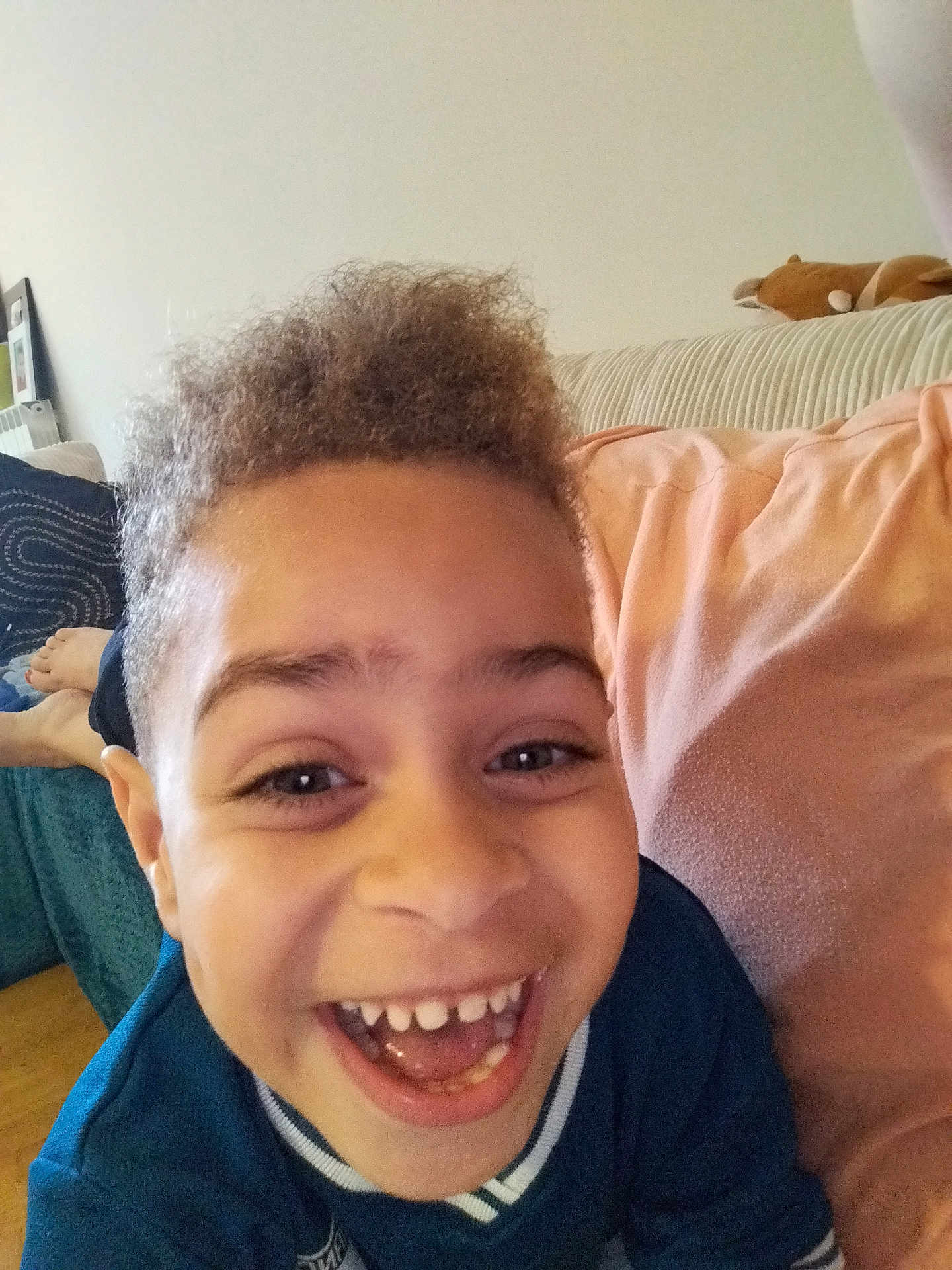 Eliott a rejoint le concours — aidez-le/la à gagner de superbes lots ! child, smiling, laughing, curly_hair, blue_shirt, couch, cushion, plush_toy, living_room, happy, face, person, indoors, closeup, cheerful, joyful, casual, relaxed, young, home
