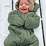baby, smiling, snow, green_snowsuit, knitted_hat, winter_clothing, outdoor, cold_weather, wooden_surface, face, happy, infant, cute, child, cozy, warm_clothing, portrait, person, winter, fun