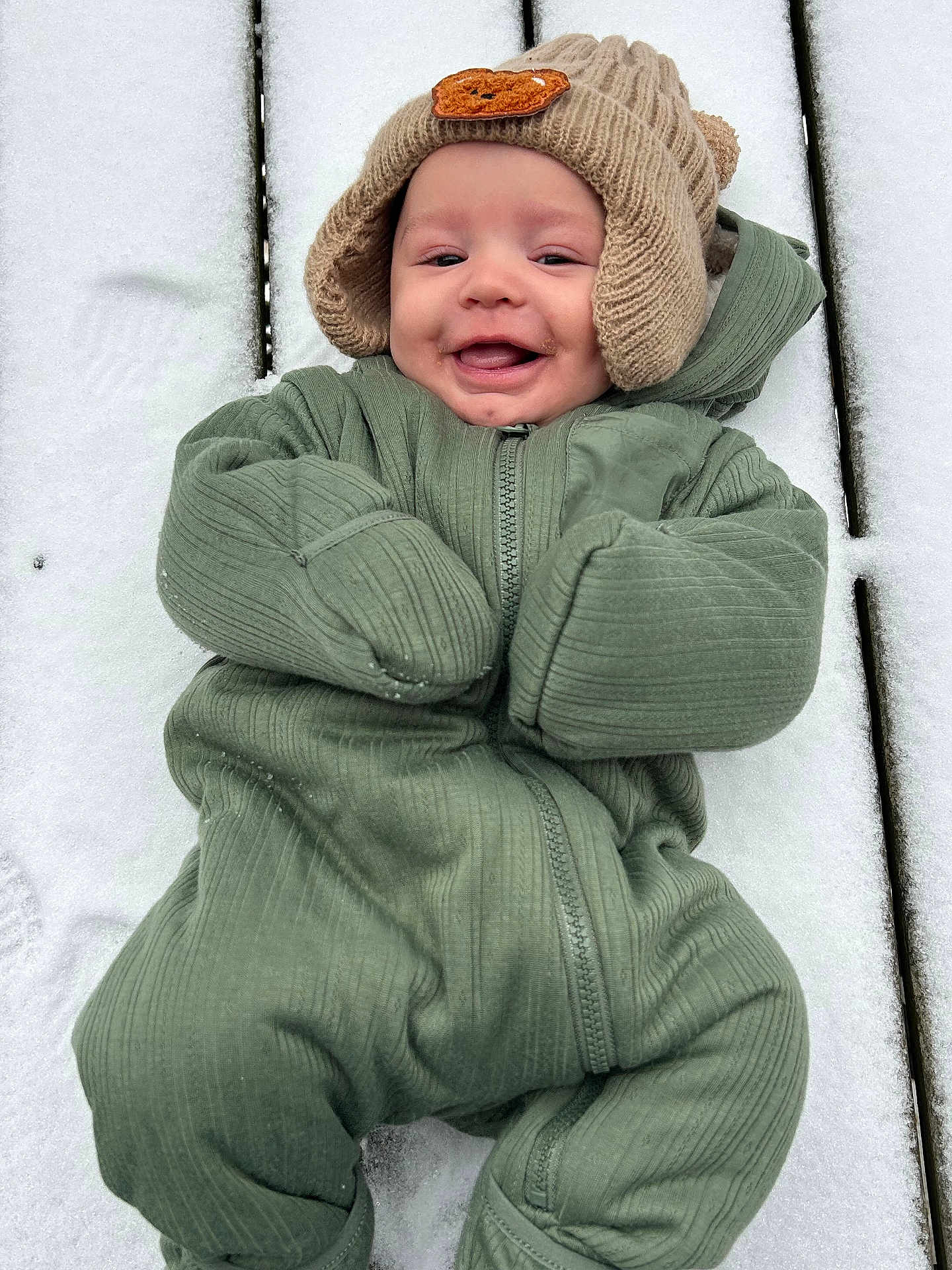 Zakari joined the competition — help win amazing prizes! baby, smiling, snow, green_snowsuit, knitted_hat, winter_clothing, outdoor, cold_weather, wooden_surface, face, happy, infant, cute, child, cozy, warm_clothing, portrait, person, winter, fun