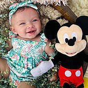 Evelyn joined the competition — help win amazing prizes! baby, child, smiling, floral_outfit, headband, plush_toy, mickey_mouse, greenery, blanket, soft_texture, happy, cute, portrait, indoor, infant, toy, colorful, sitting, person, cozy