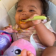 Nëylla participe au concours pour gagner de l'argent avec cette photo : baby, child, feeding, spoon, messy_face, highchair, toy, plush_toy, foot, happy, smiling, curly_hair, clothing, infant, indoor, seat, colorful, cute, person, young_child