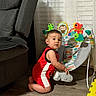 child, toddler, baby, indoor, portrait, kneeling, sitting, curious, face, short_hair, red_outfit, sportswear, activity_gym, baby_toys, frog_toy, monkey_toy, parrot_toy, turtle_toy, curtain, armchair