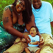 Adrian joined the competition — help win amazing prizes! baby, smiling, family, outdoor, grass, wildflowers, happy, sitting, adult, person, casual_clothing, glasses, tattoo, green_dress, shorts, white_shirt, barefoot, joy, nature, portrait