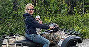 Xander is registered to the contest to win money with this photo: adult, baby, atv, all_terrain_vehicle, forest, outdoors, evergreen_trees, nature, sunlight, family, parent, infant, vehicle, motorized_vehicle, off_road, gravel, smiling, sunglasses, protective_clothing, adventure
