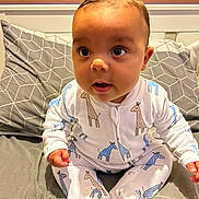 Freddie joined the competition — help win amazing prizes! baby, infant, onesie, giraffe_pattern, bed, pillow, gray, geometric_pattern, wooden_blinds, curious, sitting, indoors, child, cute, face, hands, footed_pajamas, expression, young, portrait