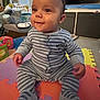 Freddie is registered to the contest to win money with this photo: baby, child, pajamas, striped_clothing, sitting, colorful_mats, foam_puzzle_mats, toys, indoor, carpet, smiling, face, person, footed_pajamas, baby_toys, storage_bins, home, play_area, cute, happy