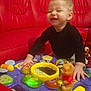 Zaïm participe au concours pour gagner de l'argent avec cette photo : toddler, child, smiling, playing, toy, activity_table, red_couch, indoor, colorful, happy, person, plastic_toy, furniture, baby, fun, young_child, caucasian, clothing, black_shirt, home