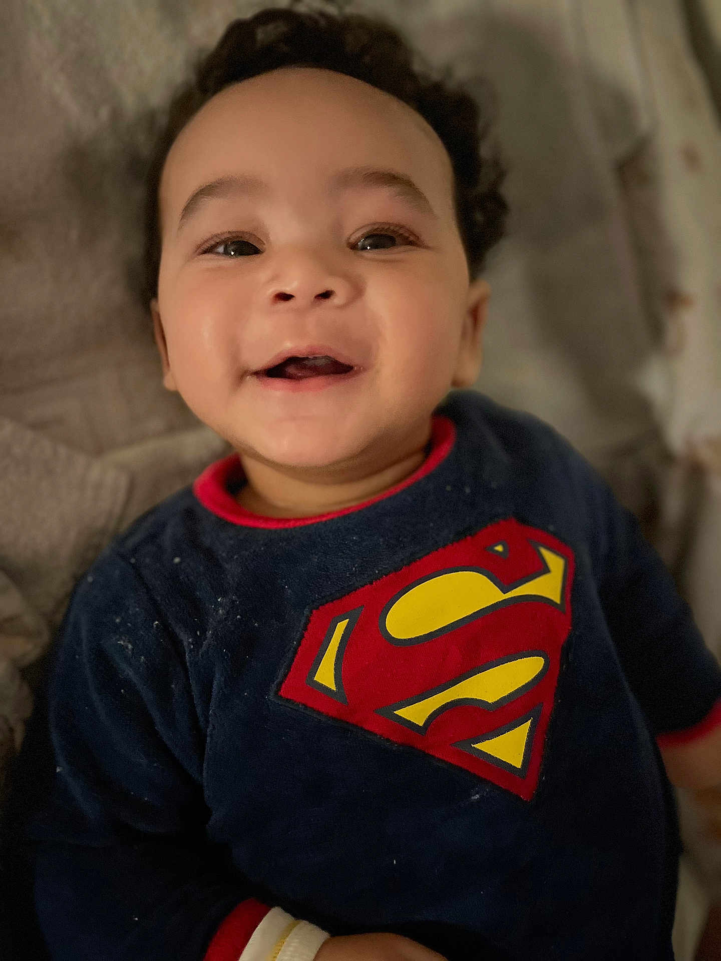 Lévi participe au concours pour gagner de l'argent avec cette photo : baby, child, superman_logo, smiling, face, curly_hair, blue_shirt, red_trim, blanket, closeup, portrait, happy, infant, cute, indoors, young_child, apparel, person, toddler, soft_lighting