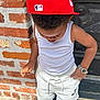toddler, child, red_cap, white_tank_top, shorts, mickey_mouse, sneakers, watch, brick_wall, door, outdoor, standing, looking_down, fashion, casual_clothing, sunlight, porch, concrete, curious, cute