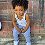 child, toddler, smiling, curly_hair, white_tank_top, gray_pants, sneakers, sitting, brick_steps, gold_necklace, earrings, outdoor, urban, metal_railing, confident, cute, portrait, person, happy, fashion