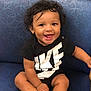 baby, child, smiling, curly_hair, black_shirt, nike, socks, blue_chair, happy, infant, cute, seated, indoor, portrait, person, young_child, comfortable, joyful, casual_clothing, adorable