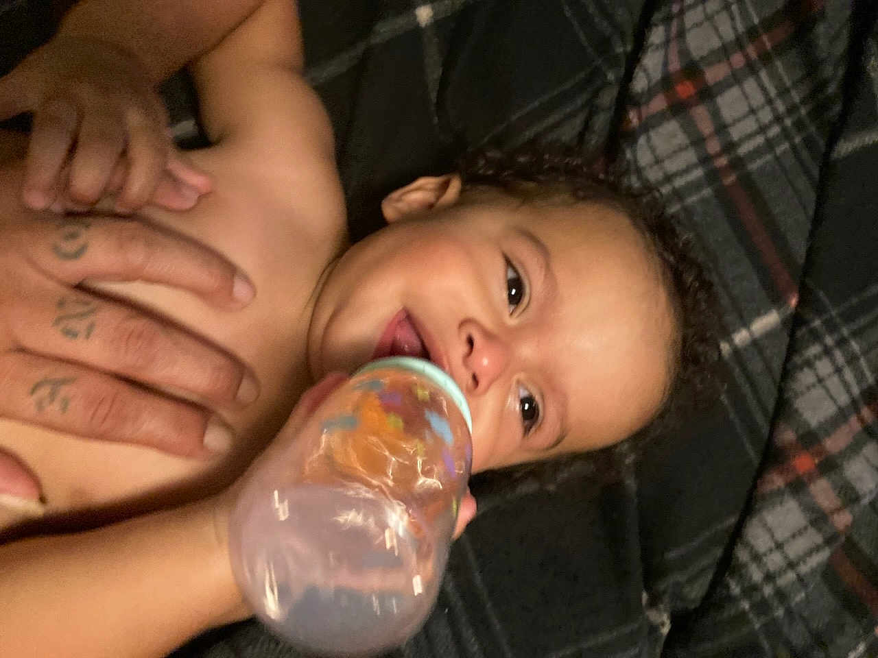 Orlando is registered to the contest to win money with this photo: baby, bottle, hand, blanket, plaid, child, drinking, smiling, person, infant, skin, cute, lying_down, happy, young, care, comfort, close_up, support, nurture