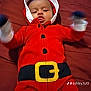 Orlando is registered to the contest to win money with this photo: baby, bed, belt, blanket, child, cute, expression, face, festive, gloves, holiday_outfit, hood, indoor, infant, lying_down, person, playful, red_clothing, tongue_out, young_child