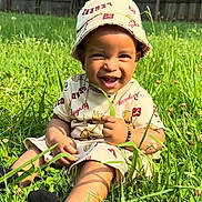 E Mon is registered to the contest to win money with this photo: toddler, child, smiling, grass, outdoor, hat, casual_clothing, sunlight, nature, greenery, happy, sitting, portrait, baby, cute, accessory, playful, daytime, field, person