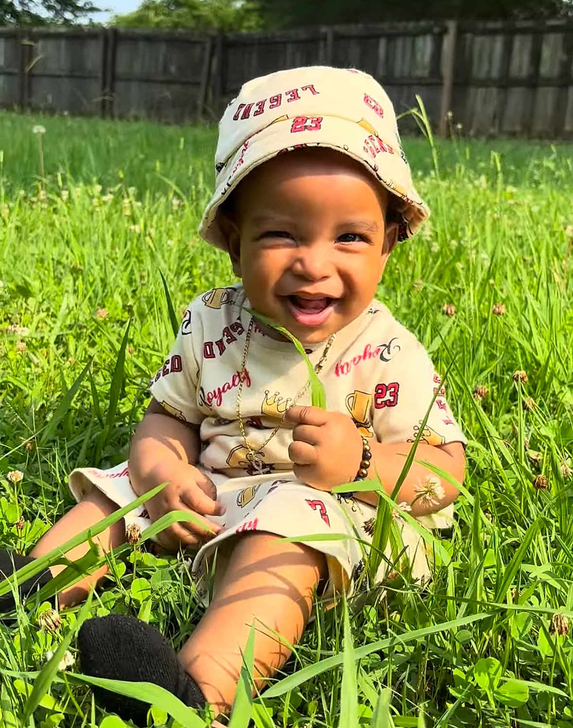 E Mon is registered to the contest to win money with this photo: toddler, child, smiling, grass, outdoor, hat, casual_clothing, sunlight, nature, greenery, happy, sitting, portrait, baby, cute, accessory, playful, daytime, field, person