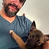 beard, blue_shirt, blur, brown_dog, candid, casual, close_up, dog, face, holding, human, indoor, interaction, man, pet, playful, portrait, puppy, smile, young_dog