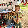 boy, dog, scout_uniform, stroller, holiday_decorations, tent, outdoor, smiling, green_shorts, brown_hair, small_dog, leaves_on_ground, gatorade_bottle, badge, patches, toy, wood, table, nature, daylight