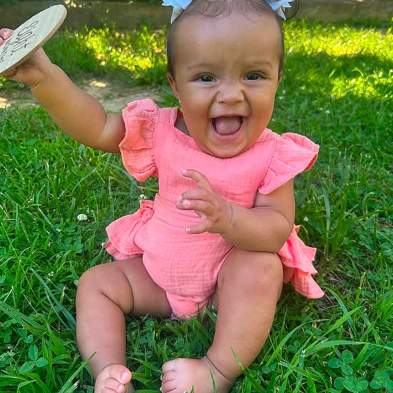 Leilani is registered to the contest to win money with this photo: baby, child, girl, smiling, laughing, grass, outdoor, greenery, hair_bows, pink_clothing, sitting, happy, cute, sunlight, nature, playful, infant, joyful, wooden_object, summer