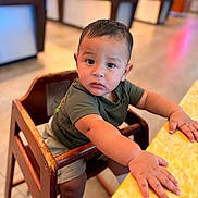 Carlos is registered to the contest to win money with this photo: toddler, child, high_chair, wooden_furniture, curious, indoor, table, yellow_table, short_hair, casual_clothing, green_shirt, beige_shorts, white_socks, small_hands, expression, face, person, photography, blurred_background, candid