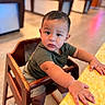 toddler, child, high_chair, wooden_furniture, curious, indoor, table, yellow_table, short_hair, casual_clothing, green_shirt, beige_shorts, white_socks, small_hands, expression, face, person, photography, blurred_background, candid