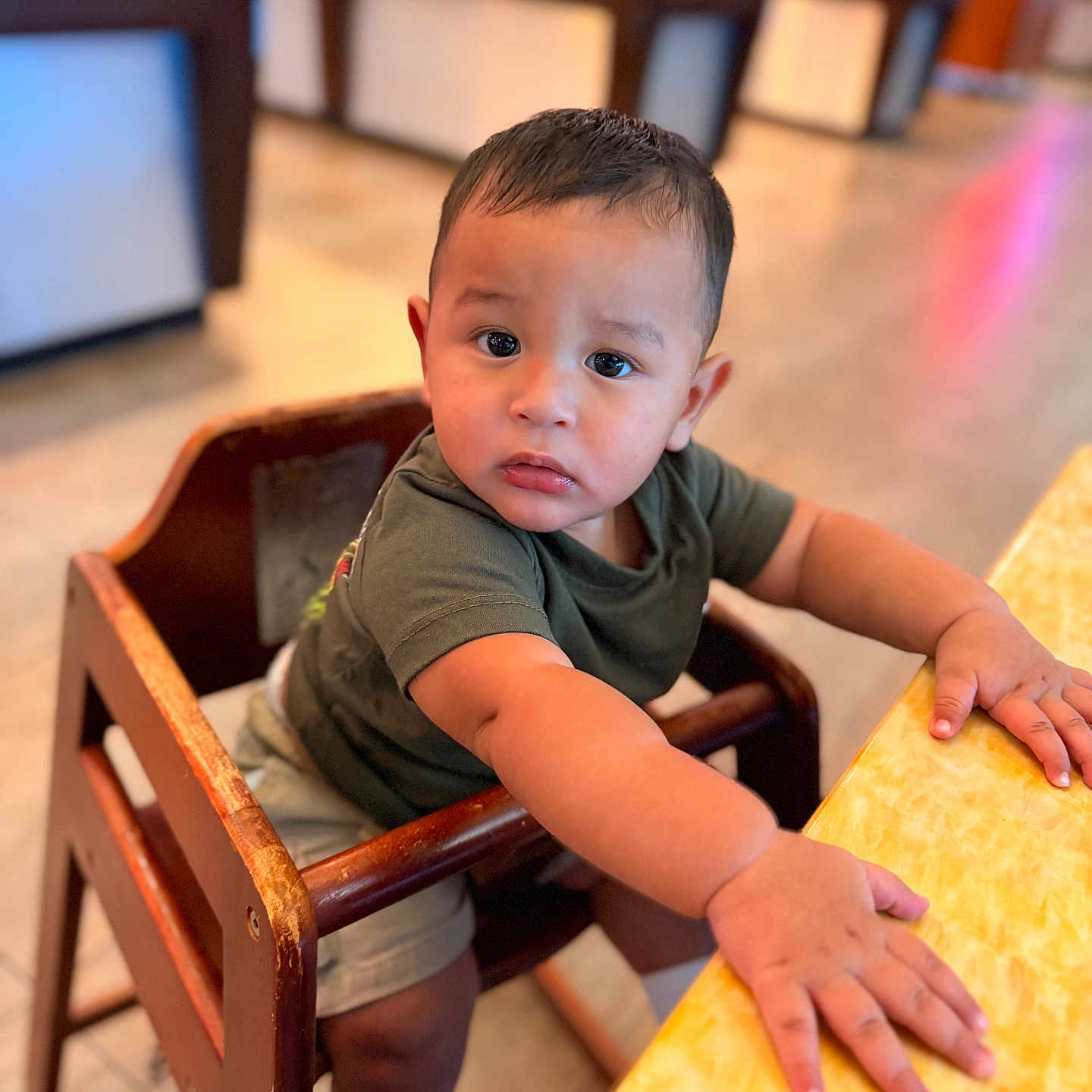 Carlos is registered to the contest to win money with this photo: toddler, child, high_chair, wooden_furniture, curious, indoor, table, yellow_table, short_hair, casual_clothing, green_shirt, beige_shorts, white_socks, small_hands, expression, face, person, photography, blurred_background, candid