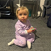 Avarose is registered to the contest to win money with this photo: toddler, child, indoor, carpet, pajamas, toy, sitting, curious, face, hairclip, furniture, photo_frame, glass_bottle, toy_truck, home, floor, sock, person, expression, living_room