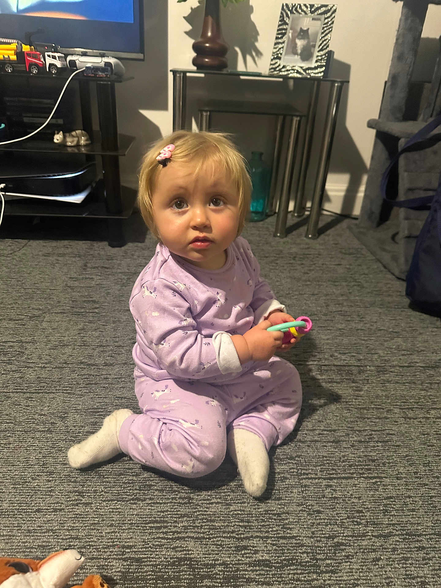 Avarose is registered to the contest to win money with this photo: toddler, child, indoor, carpet, pajamas, toy, sitting, curious, face, hairclip, furniture, photo_frame, glass_bottle, toy_truck, home, floor, sock, person, expression, living_room